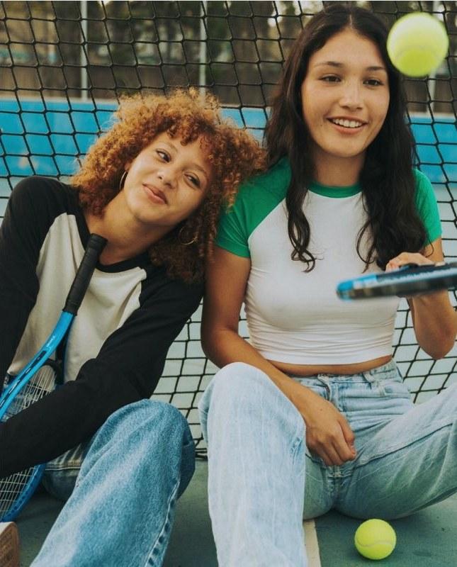 Two friends sitting against a tennis-court net with a racquet, one tossing a tennis ball